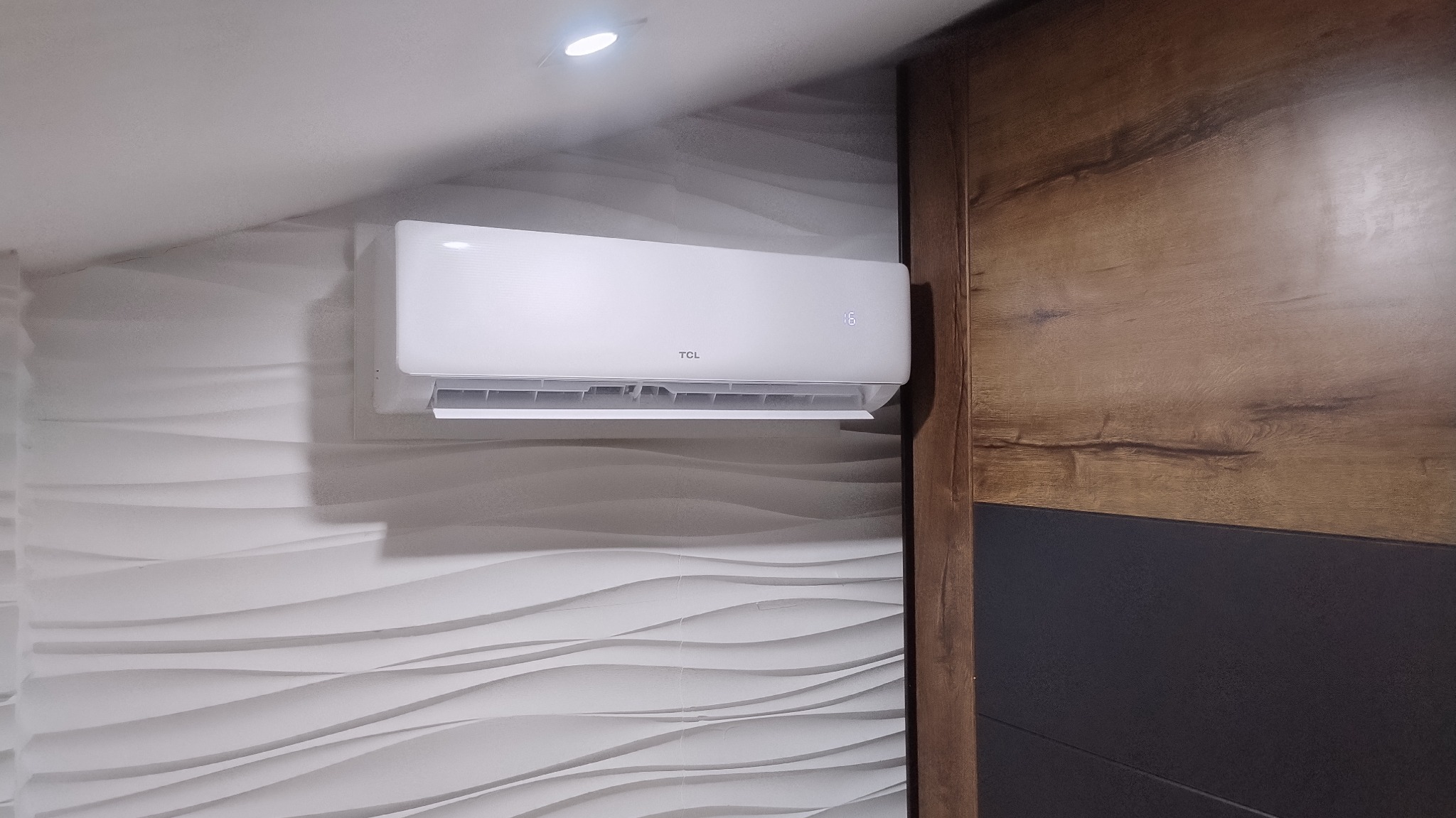 Biała klimatyzacja TCL na ścianie z dekoracyjnymi panelami 3D, widoczny fragment szafy z drewnopodobnym frontem. Nowoczesne wnętrze, oświetlenie sufitowe.