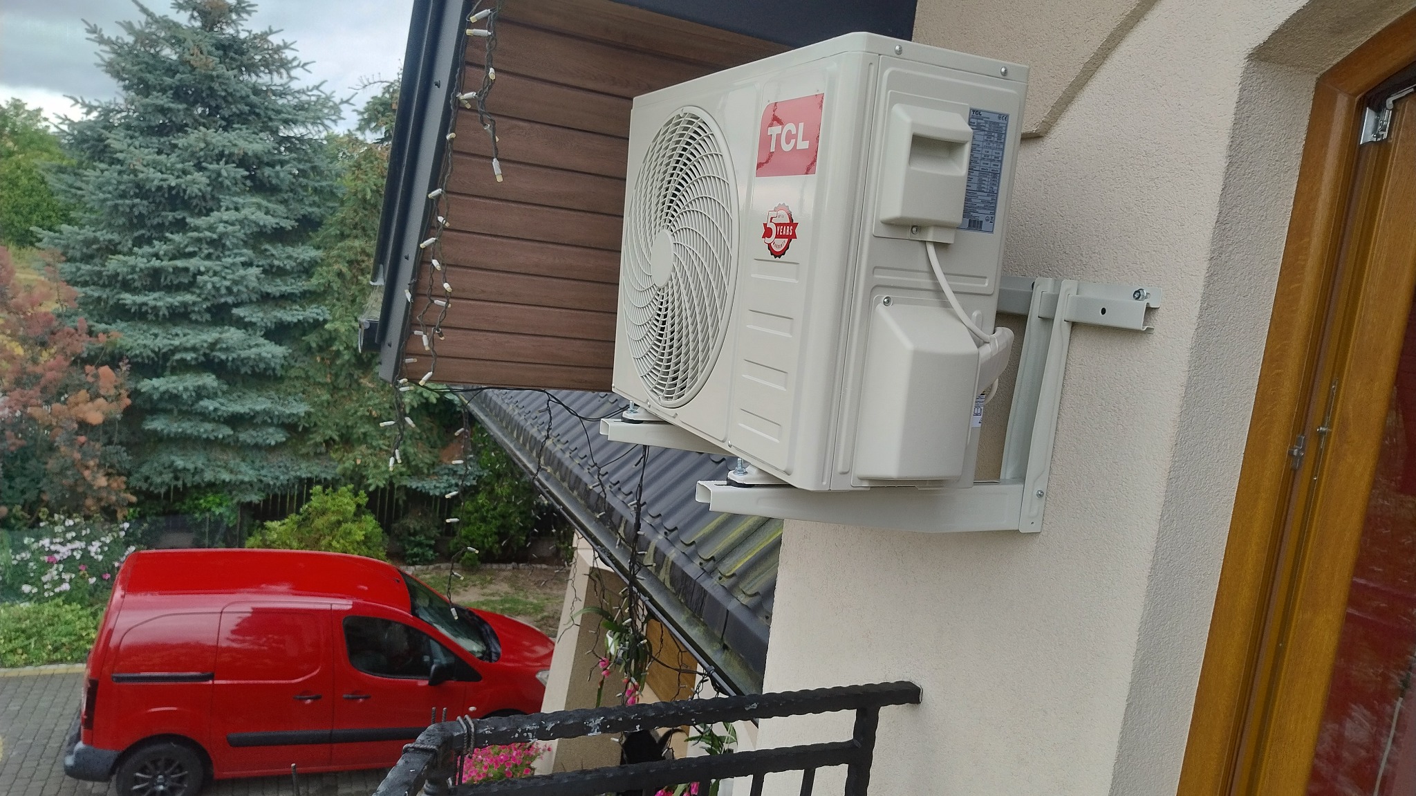 Zewnętrzna jednostka klimatyzacji TCL zamontowana na ścianie budynku, widoczny czerwony samochód dostawczy w tle. Detal montażu na wspornikach.