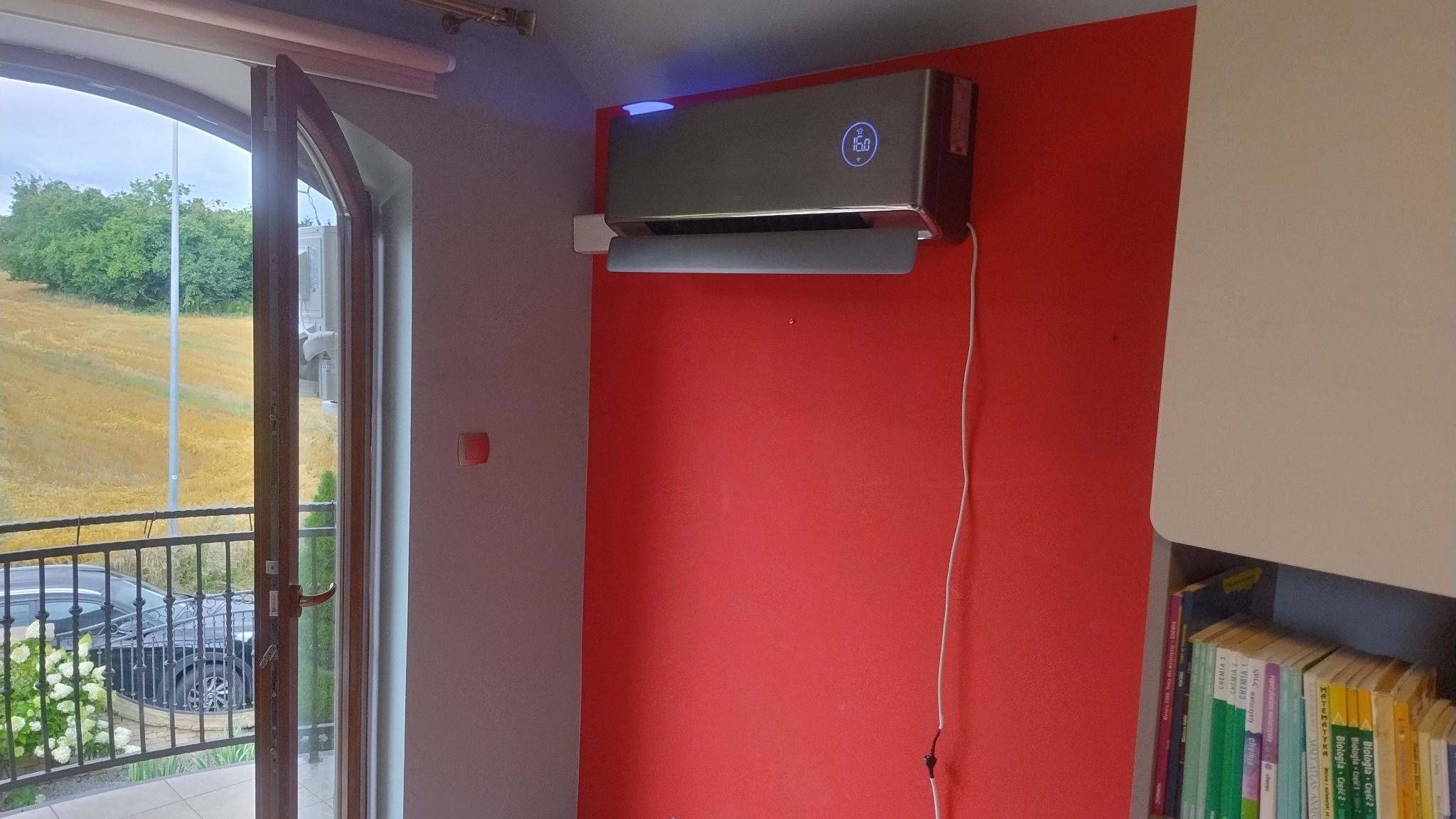 Nowoczesny klimatyzator na czerwonej ścianie w pokoju z otwartym balkonem. Widok na pole i drzewa. Na urządzeniu wyświetla się temperatura 16 stopni.
