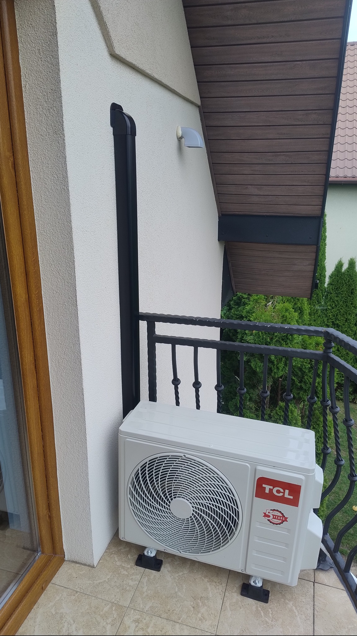 Biały klimatyzator TCL na balkonie z czarną rurą odprowadzającą i ozdobnymi barierkami; widok z boku, jasne tło budynku i zieleni.