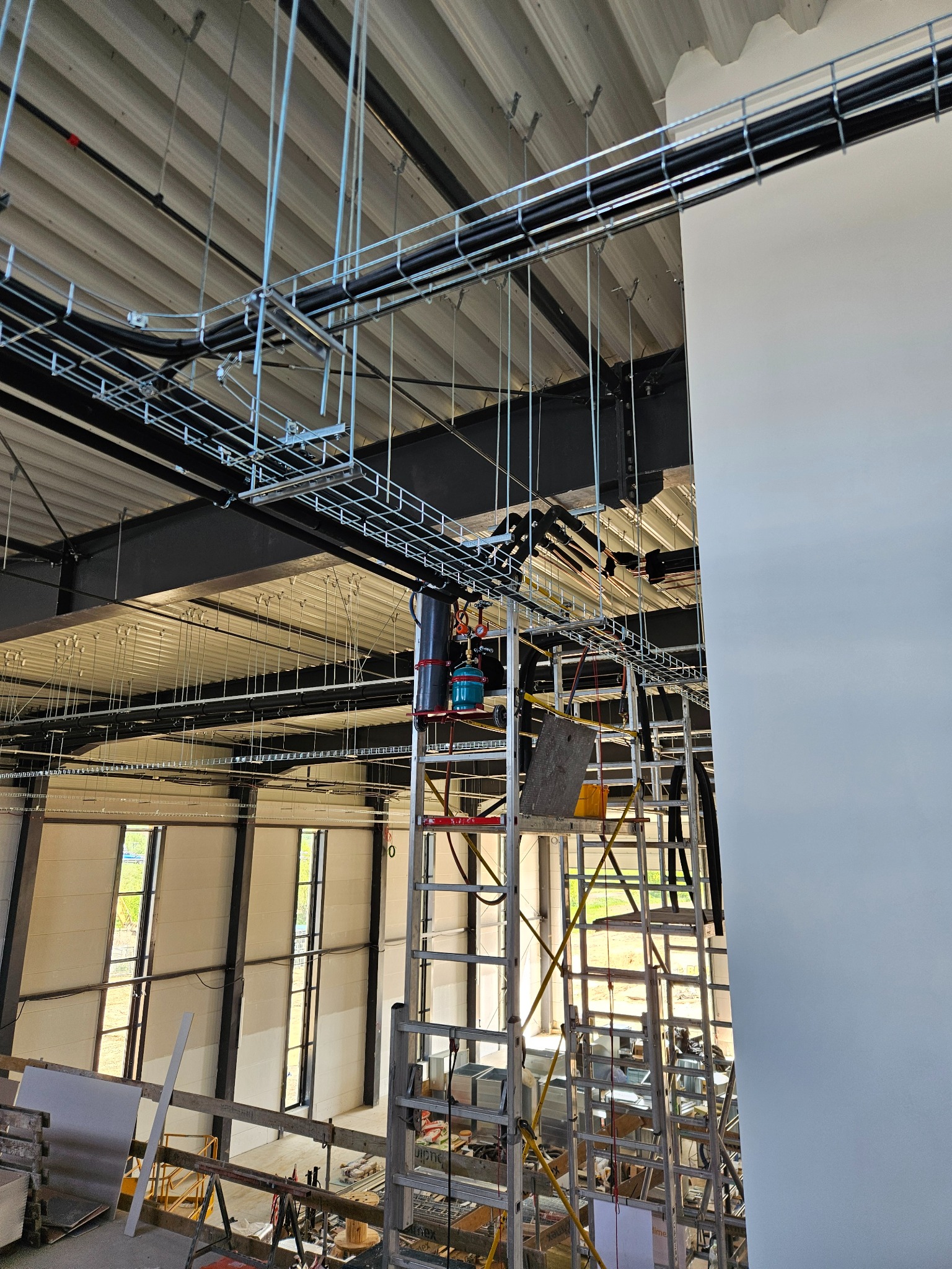 Montaż tras kablowych i rur wentylacyjnych pod sufitem w obiekcie komercyjnym w Polanowicach. Metalowe konstrukcje, drabina montażowa, surowy stan wykończenia.