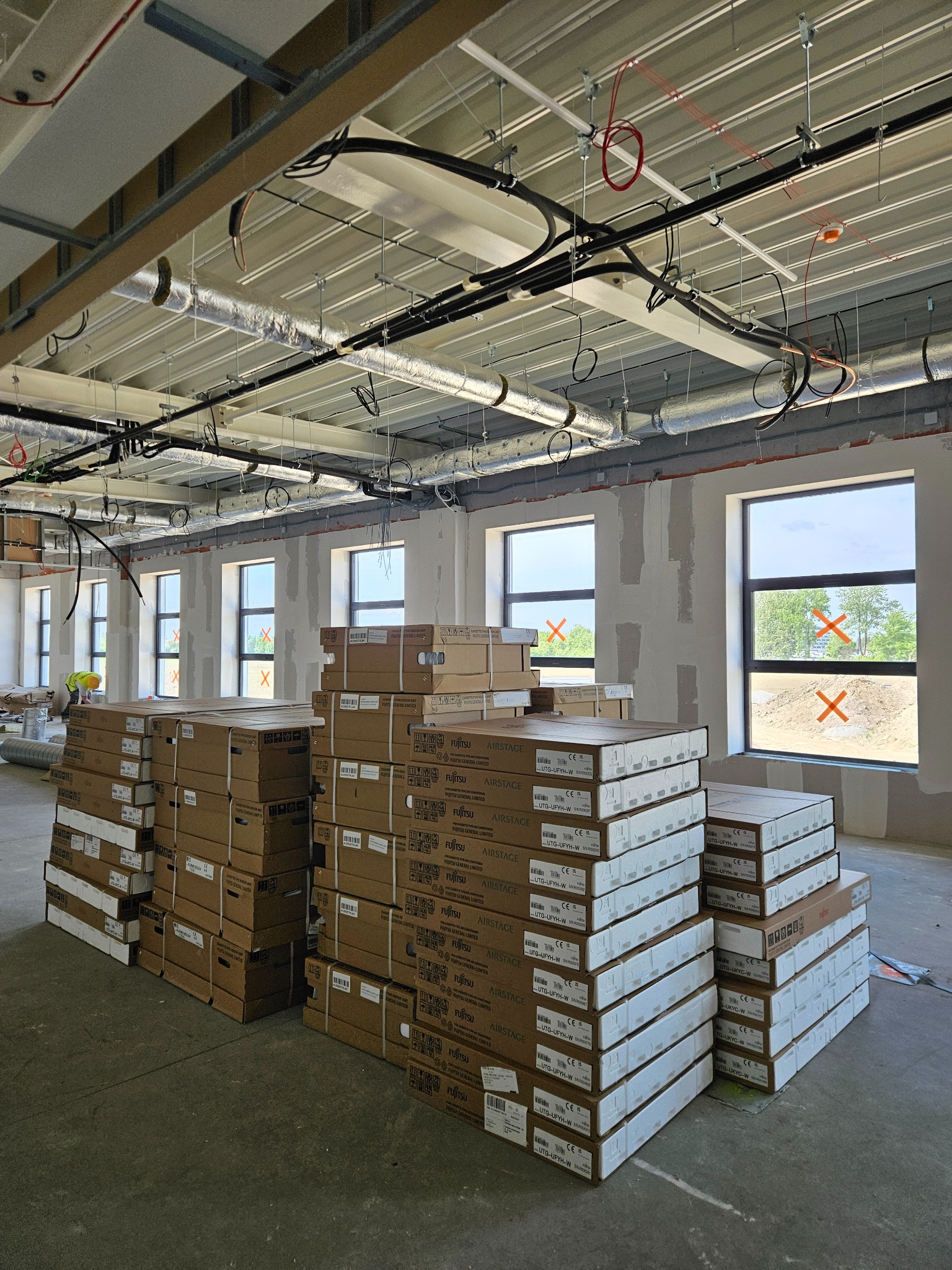 Stosy kartonów klimatyzacji Fujitsu Airstage w surowym wnętrzu biurowym, widoczne okna z taśmą zabezpieczającą i instalacje pod sufitem. Etap przed montażem.