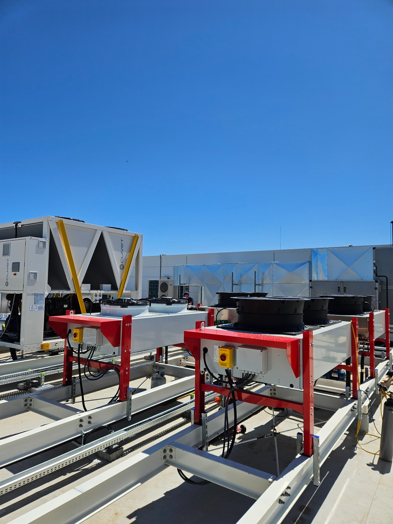 Instalacja klimatyzacji przemysłowej Clivet na dachu budynku, widoczne agregaty chłodnicze na stalowej konstrukcji, błękitne niebo w tle.