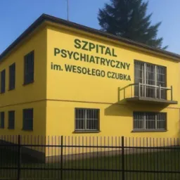 Żółty budynek szpitala psychiatrycznego z napisem 'Szpital Psychiatryczny im. Wesołego Czubka', okna z kratami i czarne metalowe ogrodzenie.