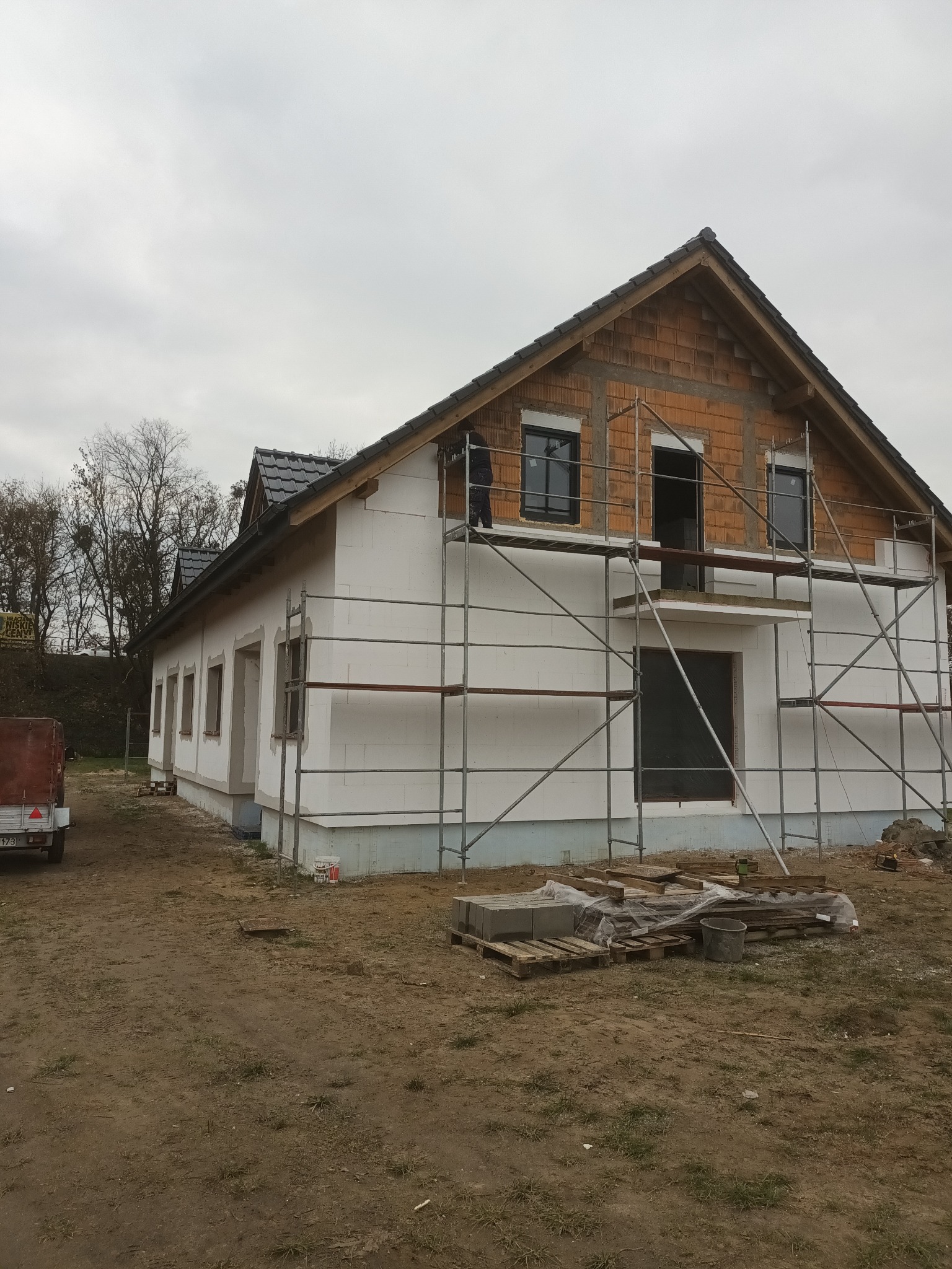 Dom w trakcie ocieplania elewacji styropianem, widoczne rusztowania i materiały budowlane. Częściowo ocieplona ściana szczytowa z cegły. Pochmurne niebo w tle.