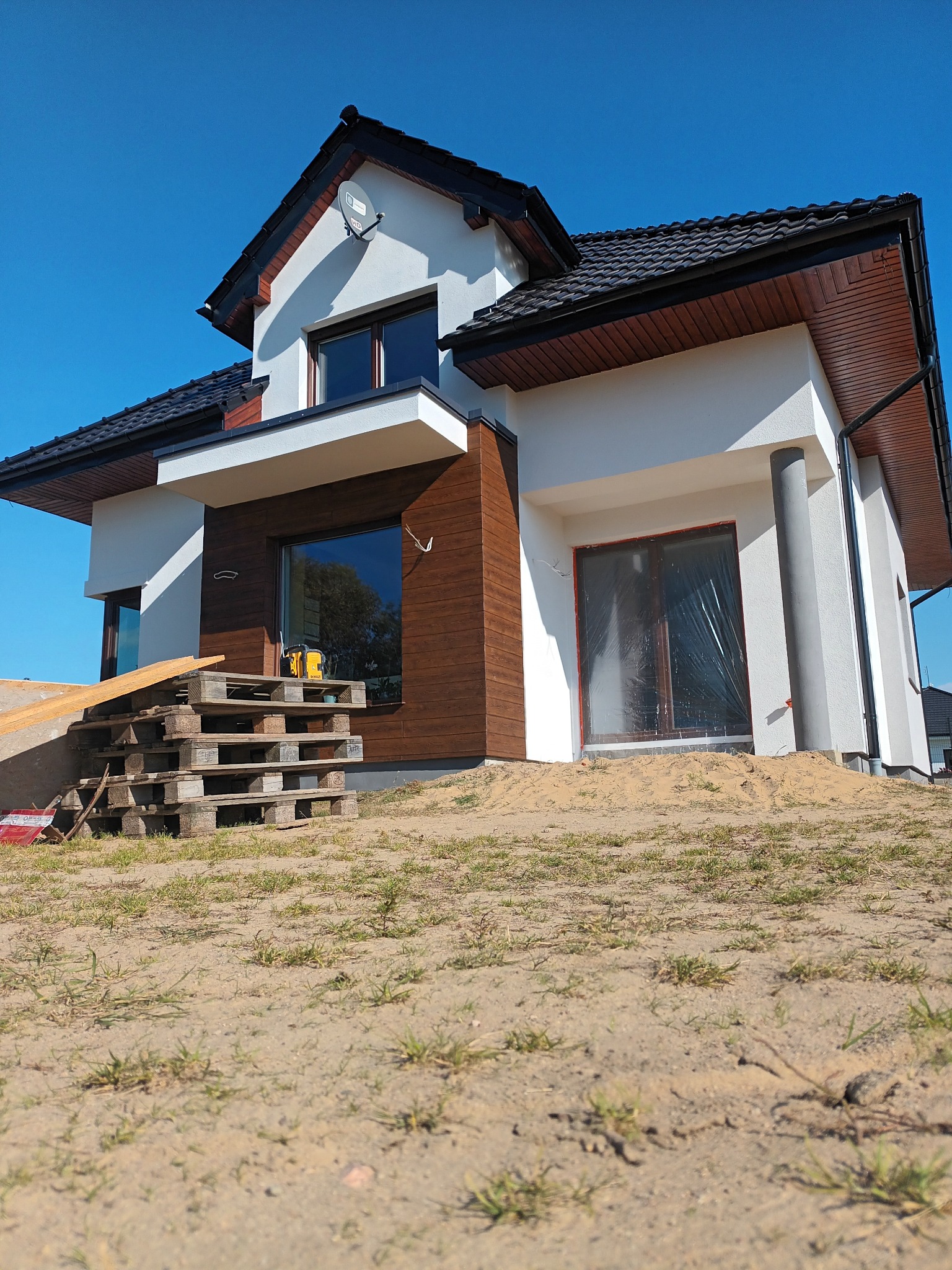 Nowoczesny dom jednorodzinny w trakcie wykończenia elewacji, z widocznymi paletami i narzędziami budowlanymi. Jasne tynki kontrastują z drewnianą okładziną.