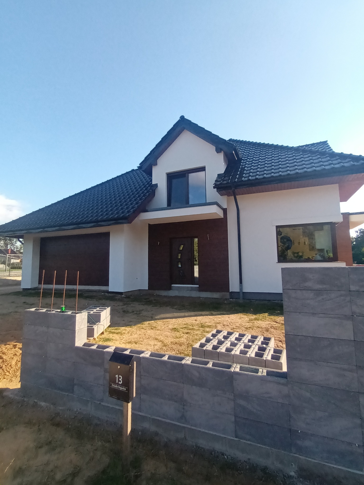 Nowoczesny dom jednorodzinny z ciemną dachówką i elementami drewnopodobnymi na elewacji, w trakcie budowy ogrodzenia z szarych bloczków betonowych.