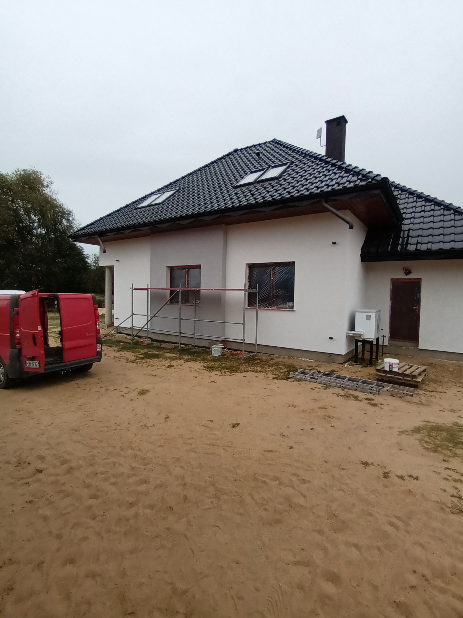 Dom jednorodzinny z czarnym dachem i białą elewacją w trakcie malowania. Widoczne rusztowania, drabina i samochód dostawczy. Okna zabezpieczone folią.