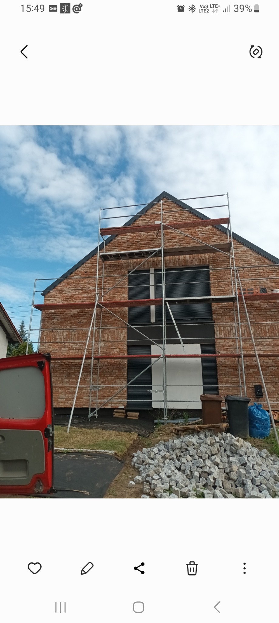 Budowa domu z cegły klinkierowej w Parkowie. Widoczne rusztowania, okna z roletami zewnętrznymi i sterta kostki brukowej. Częściowo widoczny czerwony samochód dostawczy.