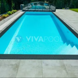 Prostokątny basen z turkusową wodą, obłożony szarymi płytami i czarną mozaiką, z częściowo zasłoniętym zadaszeniem w tle ogrodu.