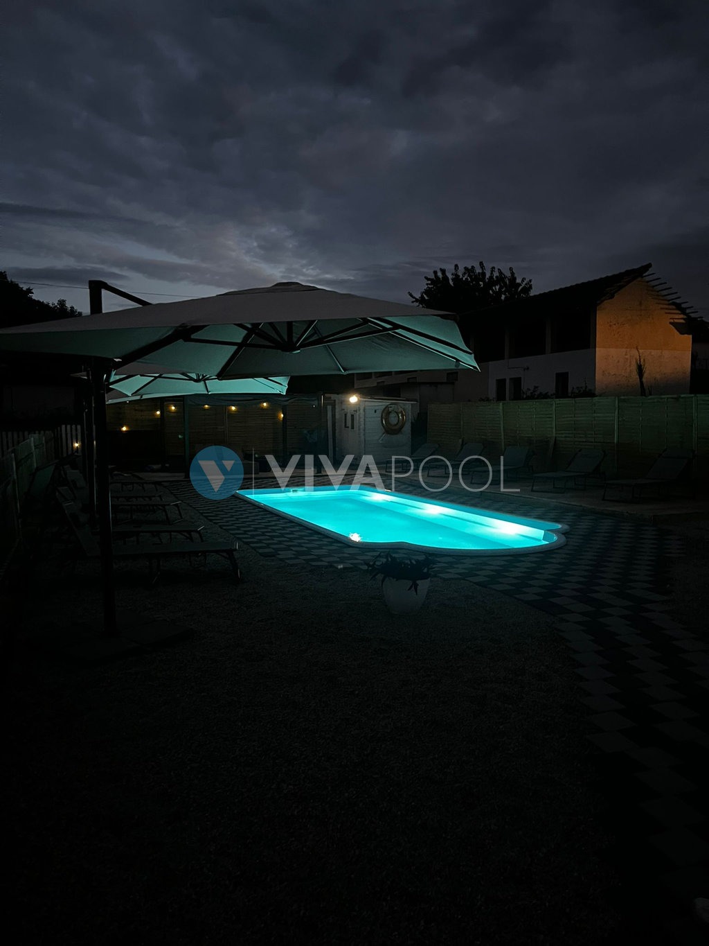 Oświetlony basen w ogrodzie nocą, zadaszony parasolem, z widocznym budynkiem w tle. Mocne oświetlenie wody tworzy kontrast z ciemnym niebem.