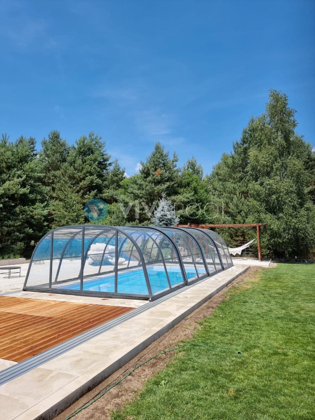 Kryty basen z przesuwnym zadaszeniem w ogrodzie, obok drewniany taras i trawnik. W tle drzewa iglaste i błękitne niebo. Hamak na drewnianej konstrukcji.