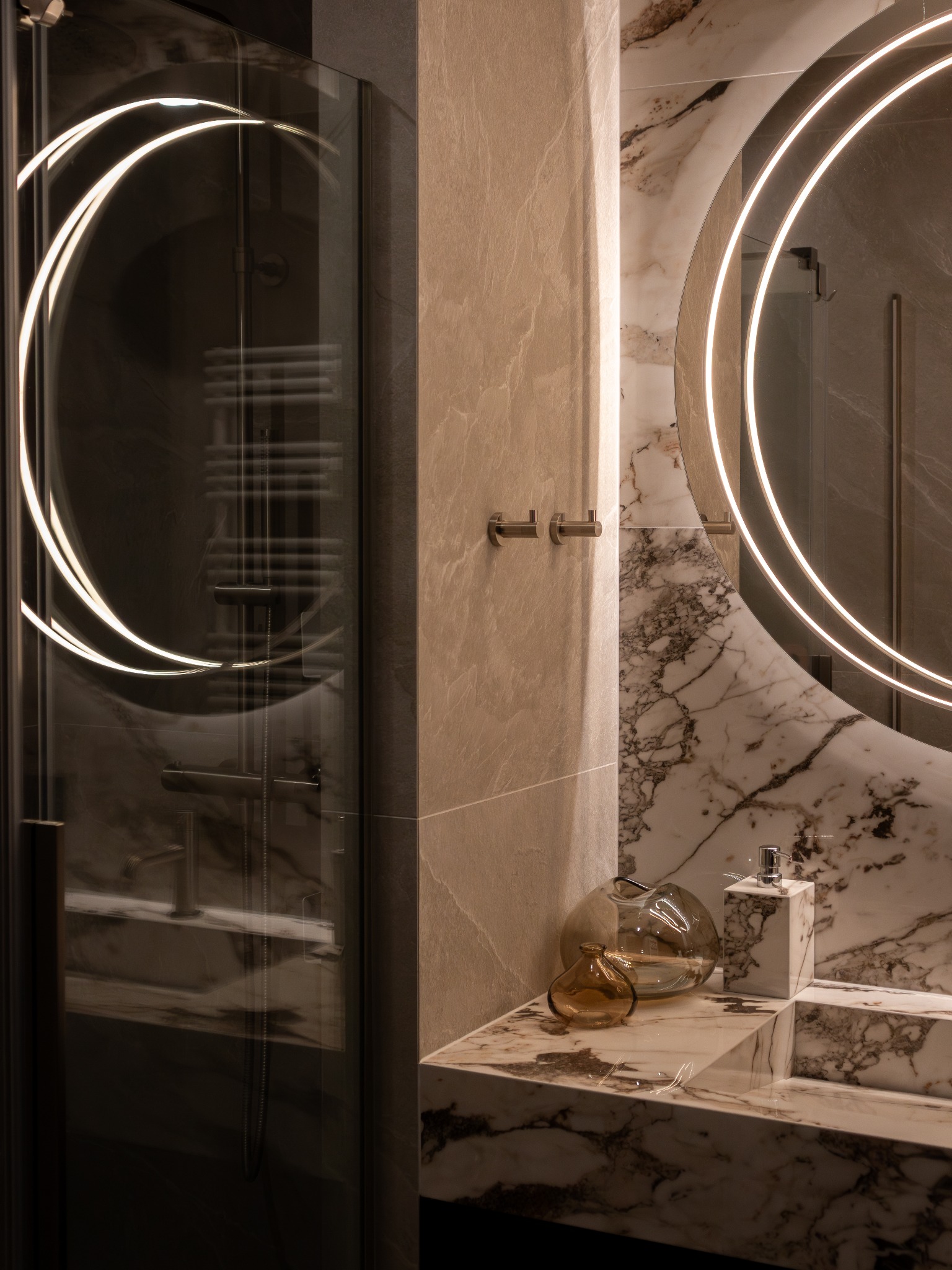 Nowoczesna łazienka: marmurowa umywalka z dozownikiem, okrągłe lustro z LED, szklana kabina prysznicowa i dekoracyjne wazony w ciepłej tonacji.