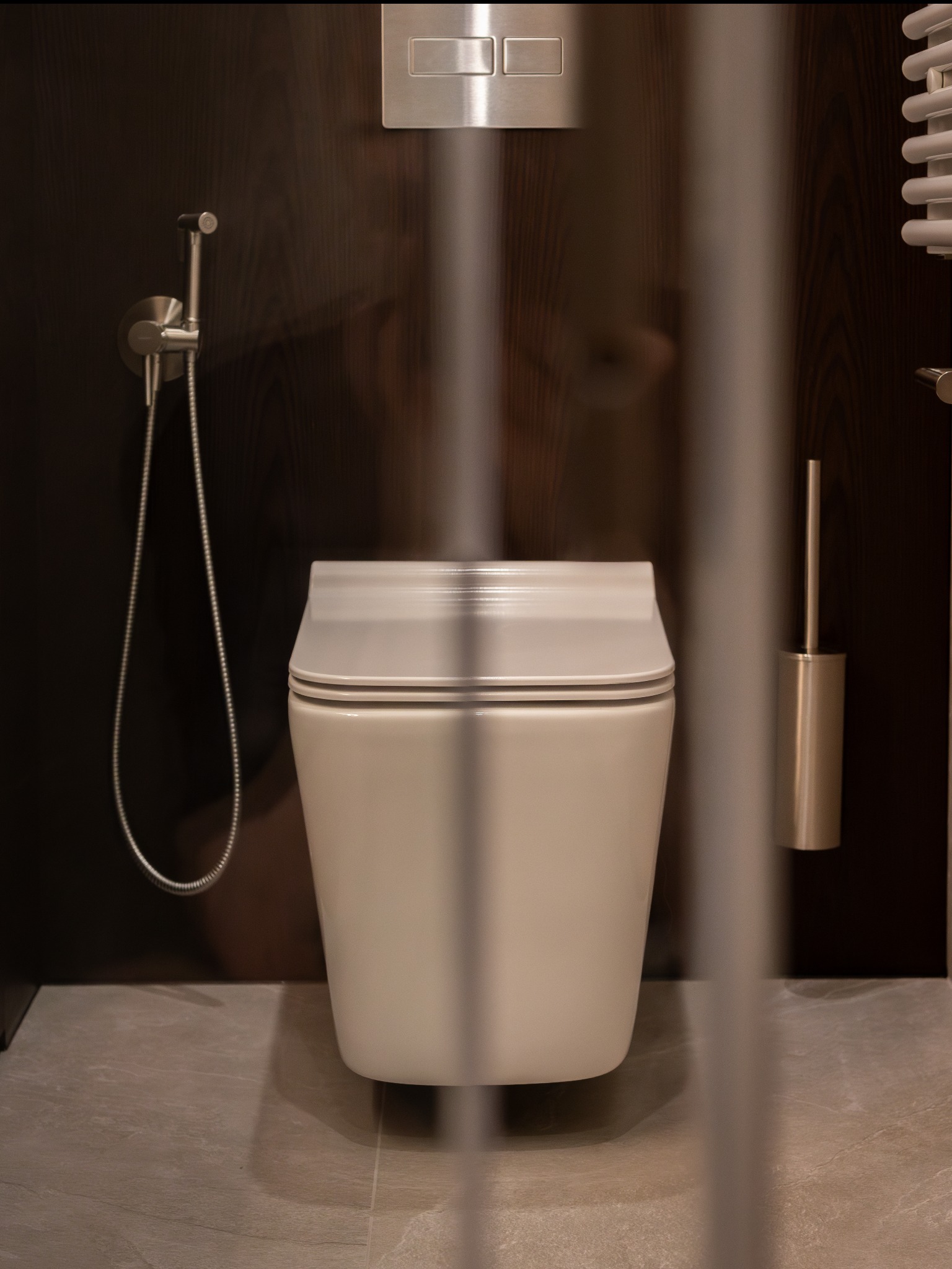 Nowoczesna, podwieszana toaleta w beżowym odcieniu, z bidetką i szczotką WC na tle ciemnego drewna. Minimalistyczny design łazienki, widok przez szklaną kabinę.