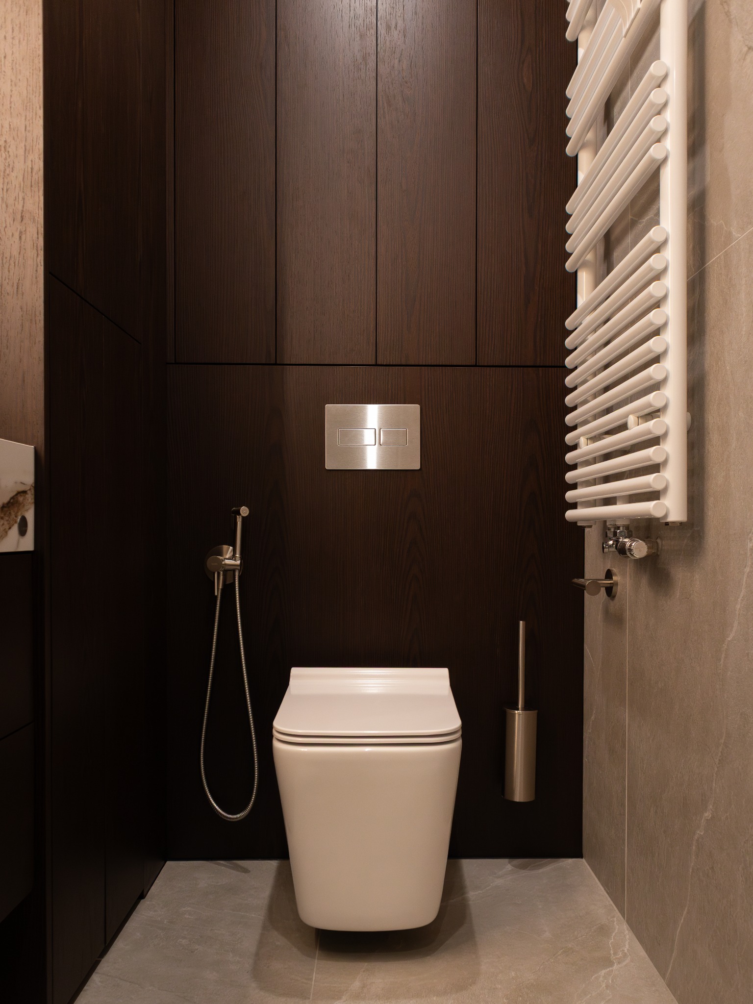 Nowoczesna łazienka z zabudową z ciemnego drewna, wiszącą toaletą i grzejnikiem drabinkowym. Minimalistyczny design z szarą podłogą i beżowymi płytkami.