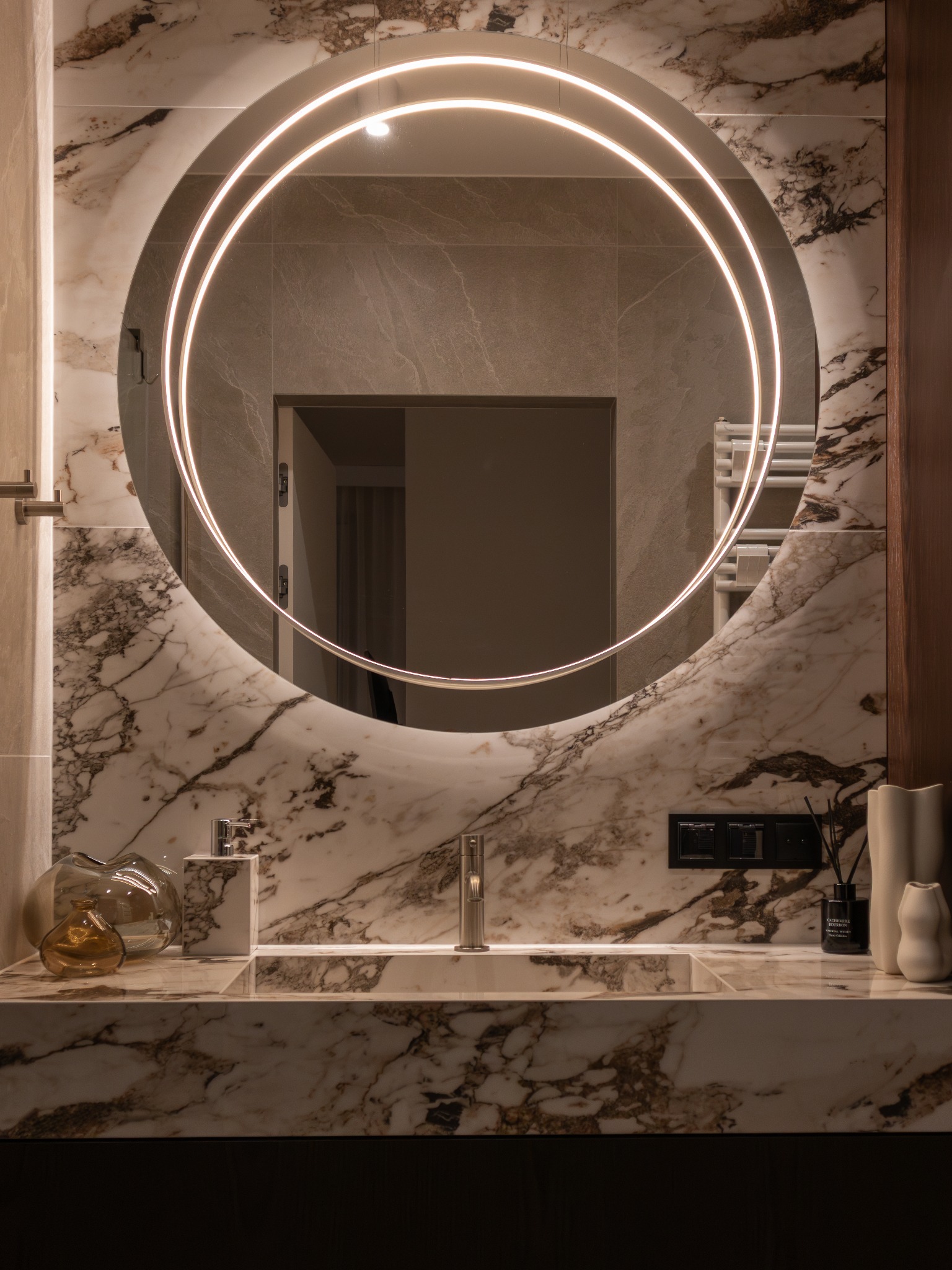 Elegancka łazienka z marmurową ścianą, okrągłym lustrem z podświetleniem LED, umywalką i dekoracyjnymi elementami. Nowoczesne wykończenie.