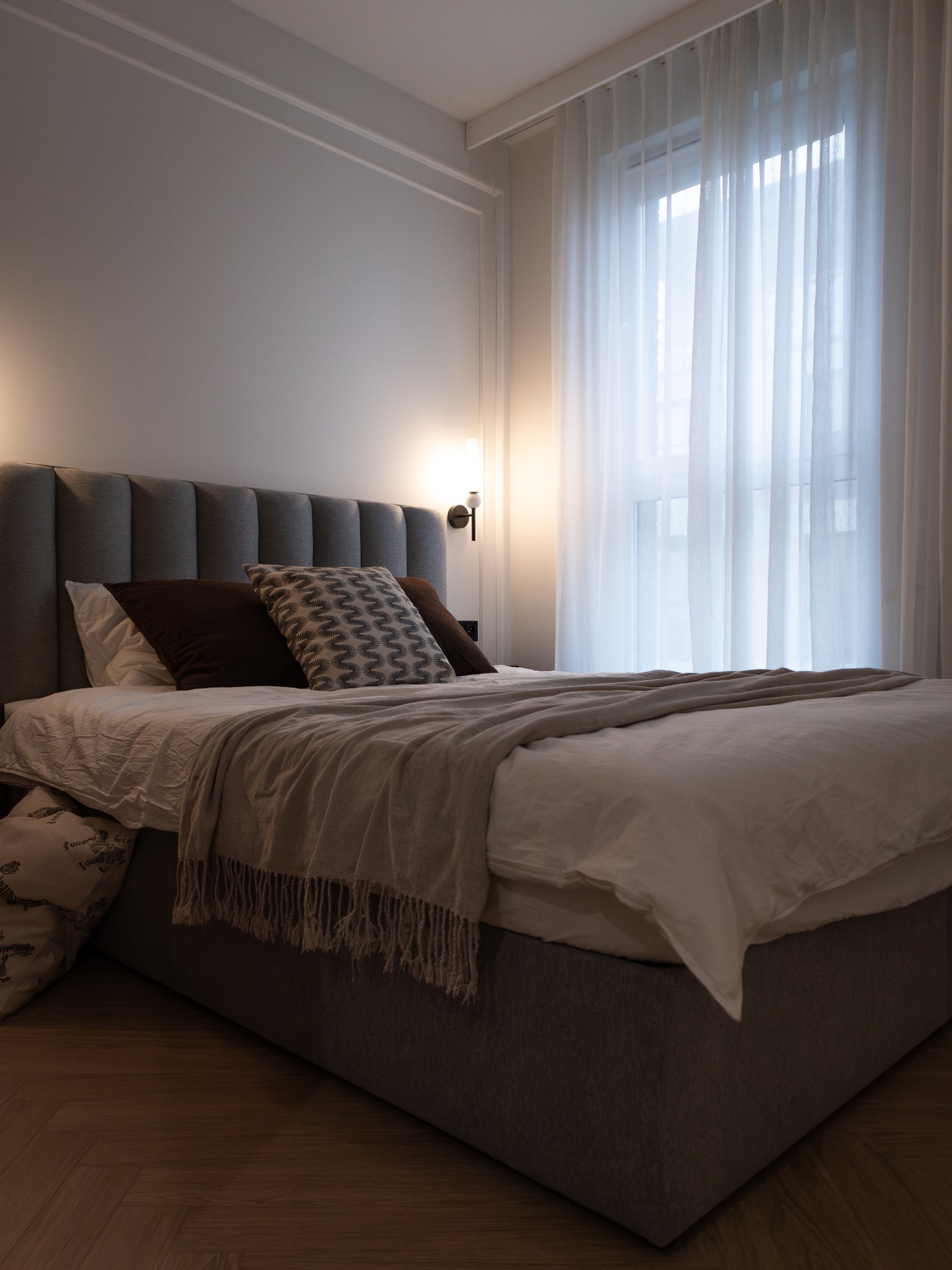 Nowoczesna sypialnia z szarym łóżkiem, wzorzystymi poduszkami i zwiewnymi, białymi zasłonami w oknie. Ciepłe światło lampki nocnej. Parkiet na podłodze.