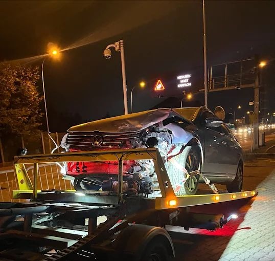 Uszkodzony samochód Volkswagen na lawecie w nocy, widoczne uszkodzenia przodu pojazdu, oświetlenie miejskie w tle.