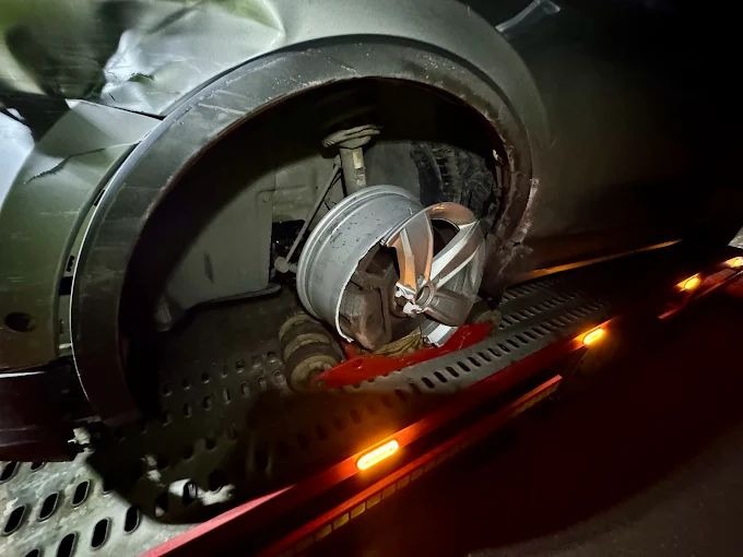 Uszkodzony samochód z urwanym kołem na platformie lawety. Widoczne wgniecenia karoserii i uszkodzone zawieszenie. Nocne oświetlenie.