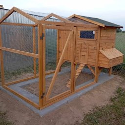 MM GROUP PODCHALE - Drewniany kurnik z siatką, osadzony na betonowej podstawie. Widoczna drabinka, okienko i budka lęgowa. Tło stanowią trawa i drzewa.