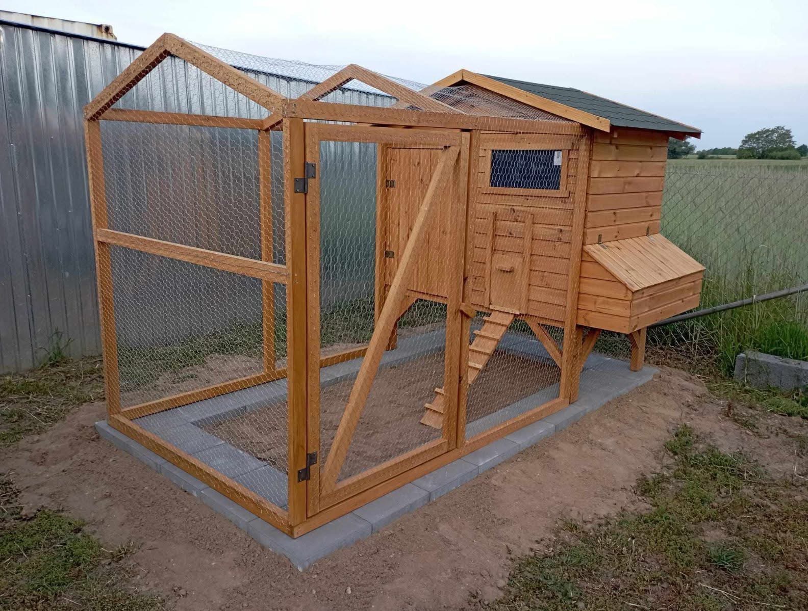 Drewniany kurnik z siatką, osadzony na betonowej podstawie. Widoczna drabinka, okienko i budka lęgowa. Tło stanowią trawa i drzewa.