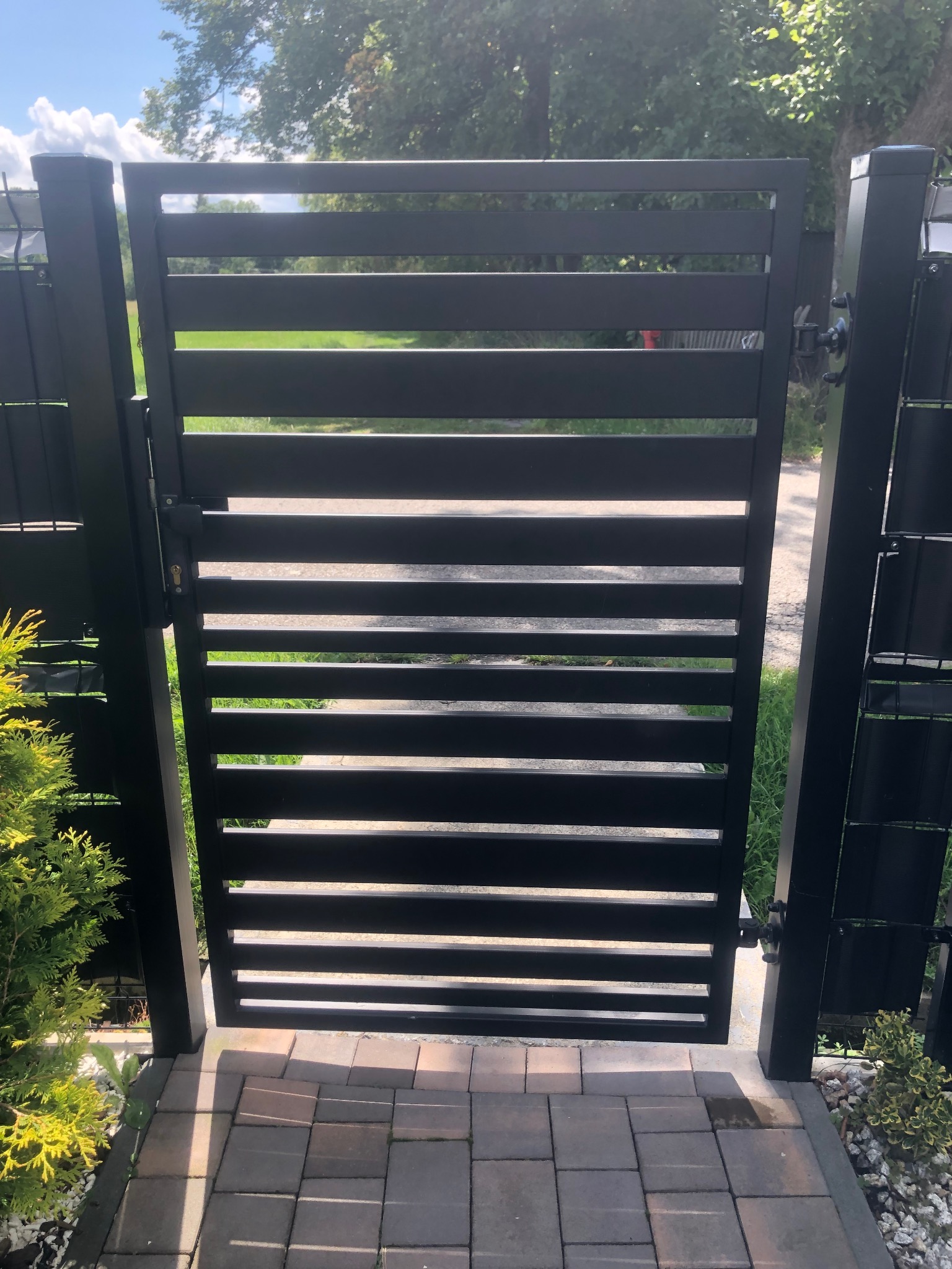 Nowoczesna, czarna brama lamelowa z zamkiem, zamontowana na posesji z kostką brukową i zielenią. Widok z bliska, w słoneczny dzień.