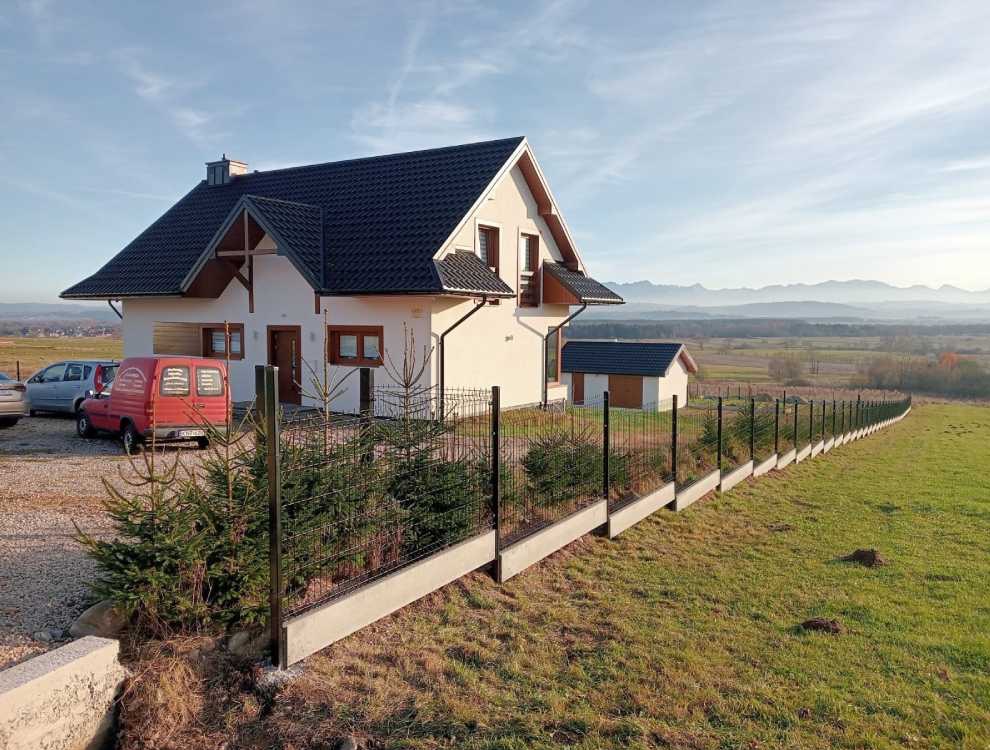 Nowoczesne ogrodzenie panelowe z podmurówką betonową, otaczające dom z ogrodem na tle górskiego krajobrazu. Widoczny czerwony samochód dostawczy.
