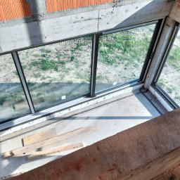 Brespol - Wnętrze surowego budynku w trakcie budowy z dużym oknem, widoczne bloczki ceramiczne i beton. Na podłodze leżą materiały budowlane.