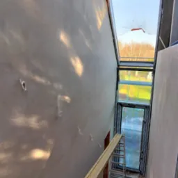 Ściana po szpachlowaniu w klatce schodowej z oknem dachowym, widok z góry. Widoczne ślady po przewodach elektrycznych i drabina. Wpadające światło słoneczne.