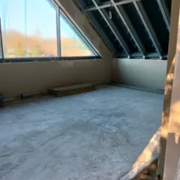 Poddasze w trakcie prac wykończeniowych: widoczne płyty karton-gips na ścianach, metalowa konstrukcja sufitu i okno dachowe zabezpieczone folią. Betonowa podłoga.