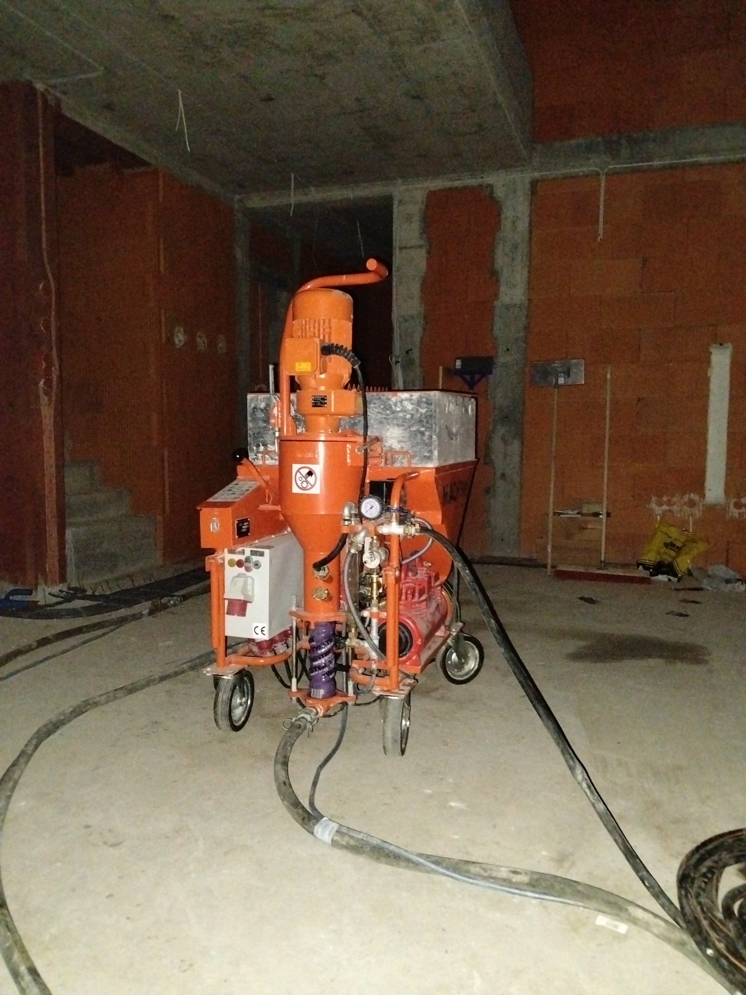 Pomarańczowa maszyna do tynkowania na kółkach w surowym wnętrzu z ceglanymi ścianami. Widoczne kable i węże maszyny. W tle niezabudowane otwory drzwiowe.