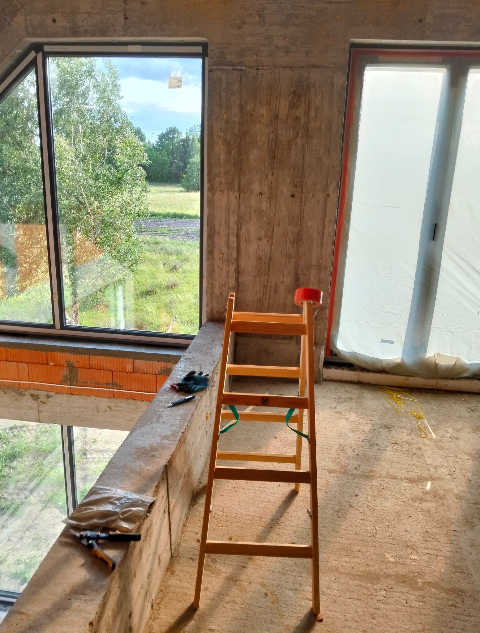 Wnętrze w trakcie prac wykończeniowych: drewniana drabina, narzędzia na surowym murze, okno z widokiem na zieleń. Jedno z okien zabezpieczone folią malarską.