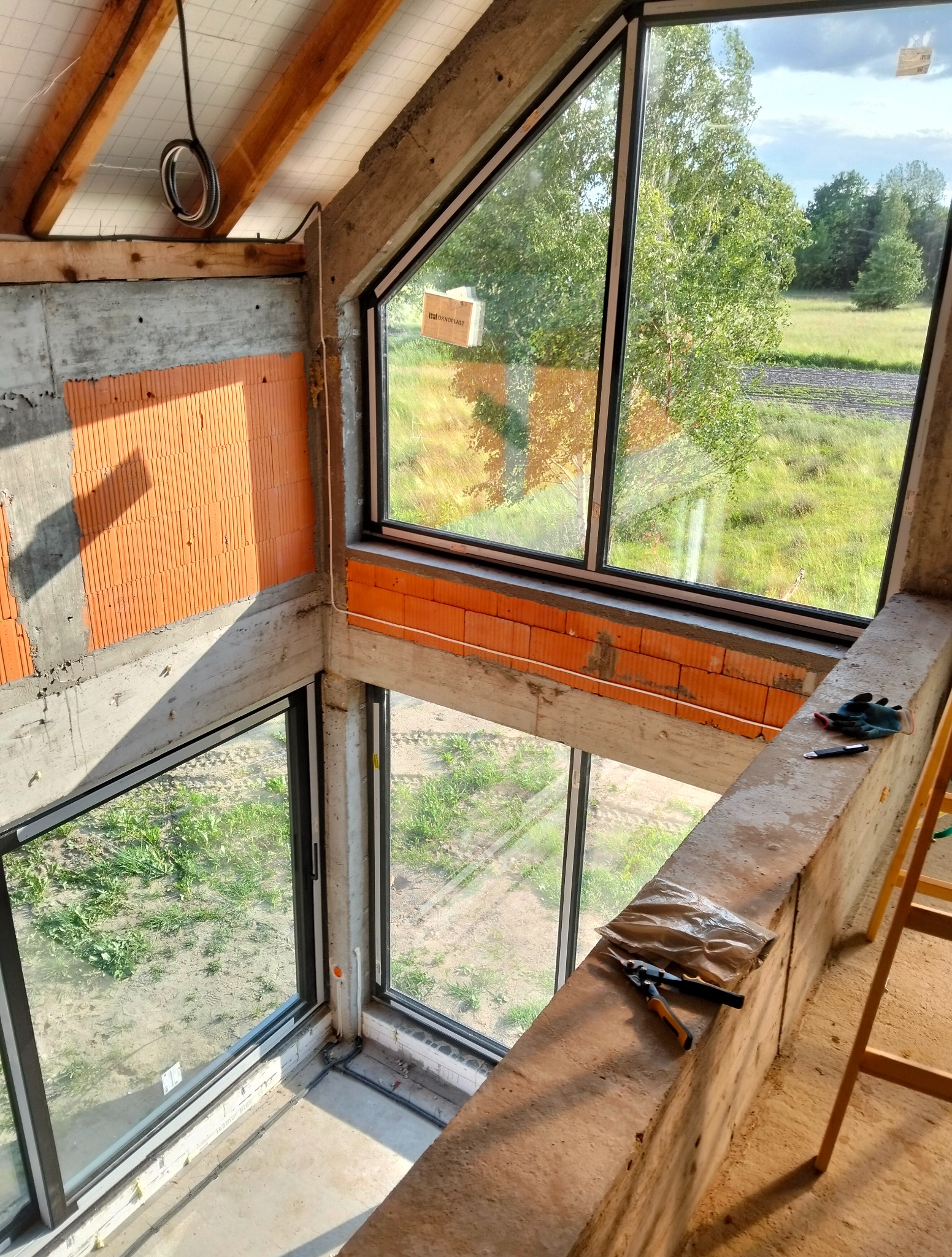 Wnętrze domu w trakcie budowy z dużymi oknami, widok na zewnątrz. Częściowo otynkowana ściana z cegły, narzędzia i drabina w kadrze. Surowy stan.