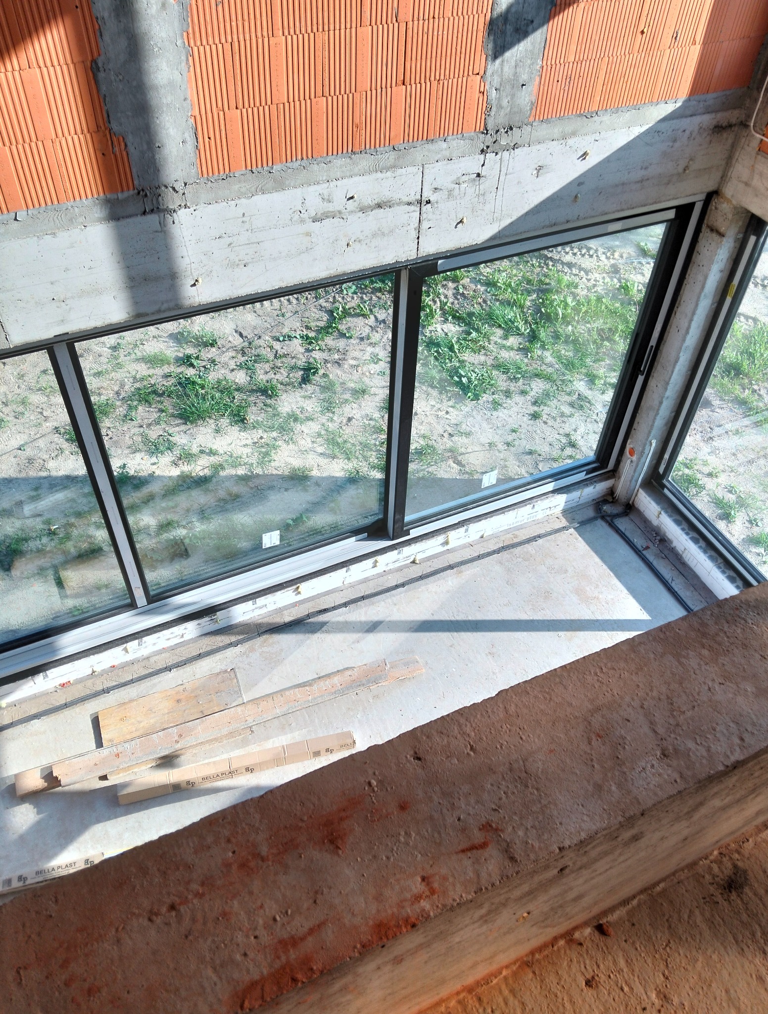 Wnętrze surowego budynku w trakcie budowy z dużym oknem, widoczne bloczki ceramiczne i beton. Na podłodze leżą materiały budowlane.