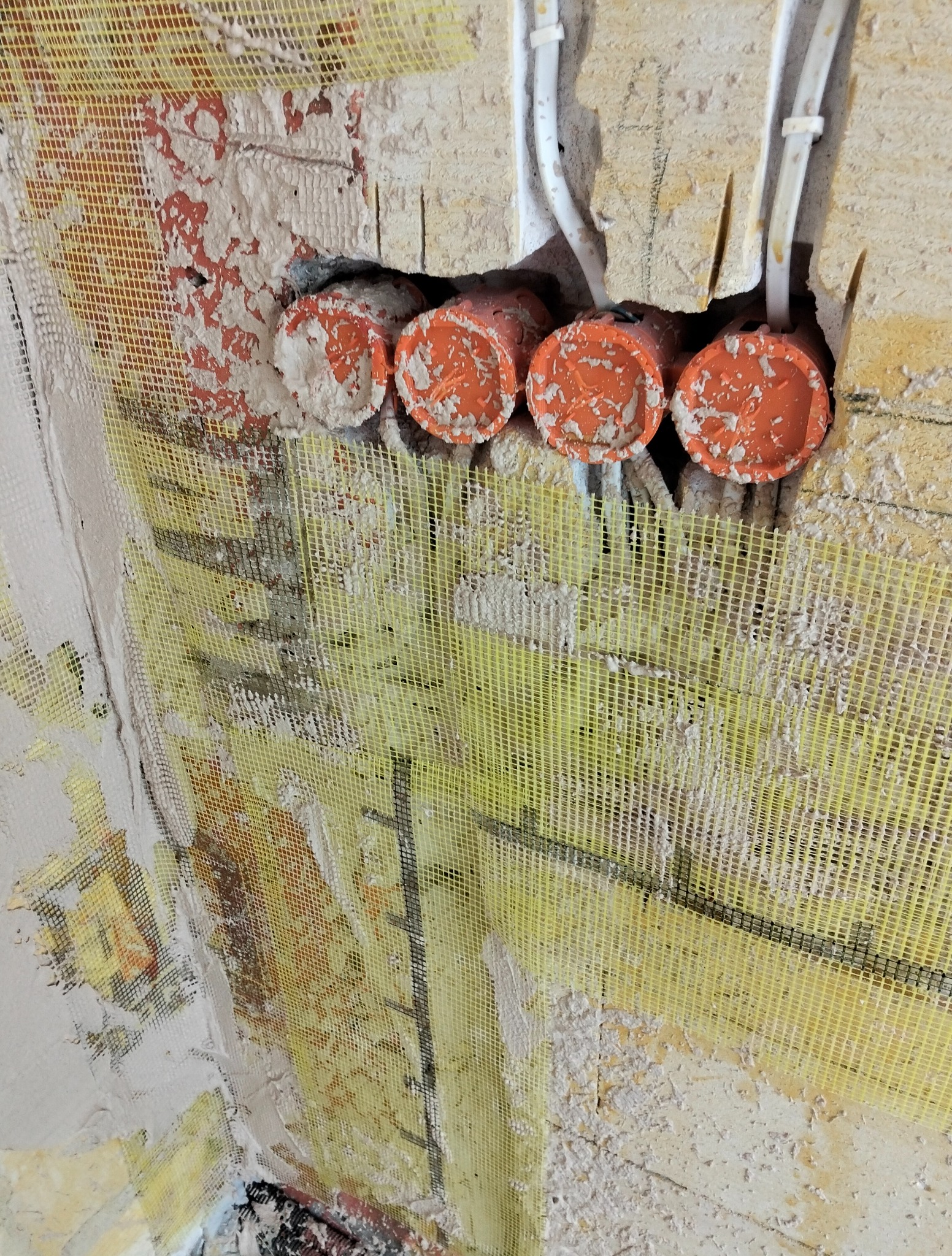 Ściana z siatką wzmacniającą, widoczne pomarańczowe rury instalacji elektrycznej i przewody. Fragment ściany przygotowany do dalszych prac wykończeniowych.