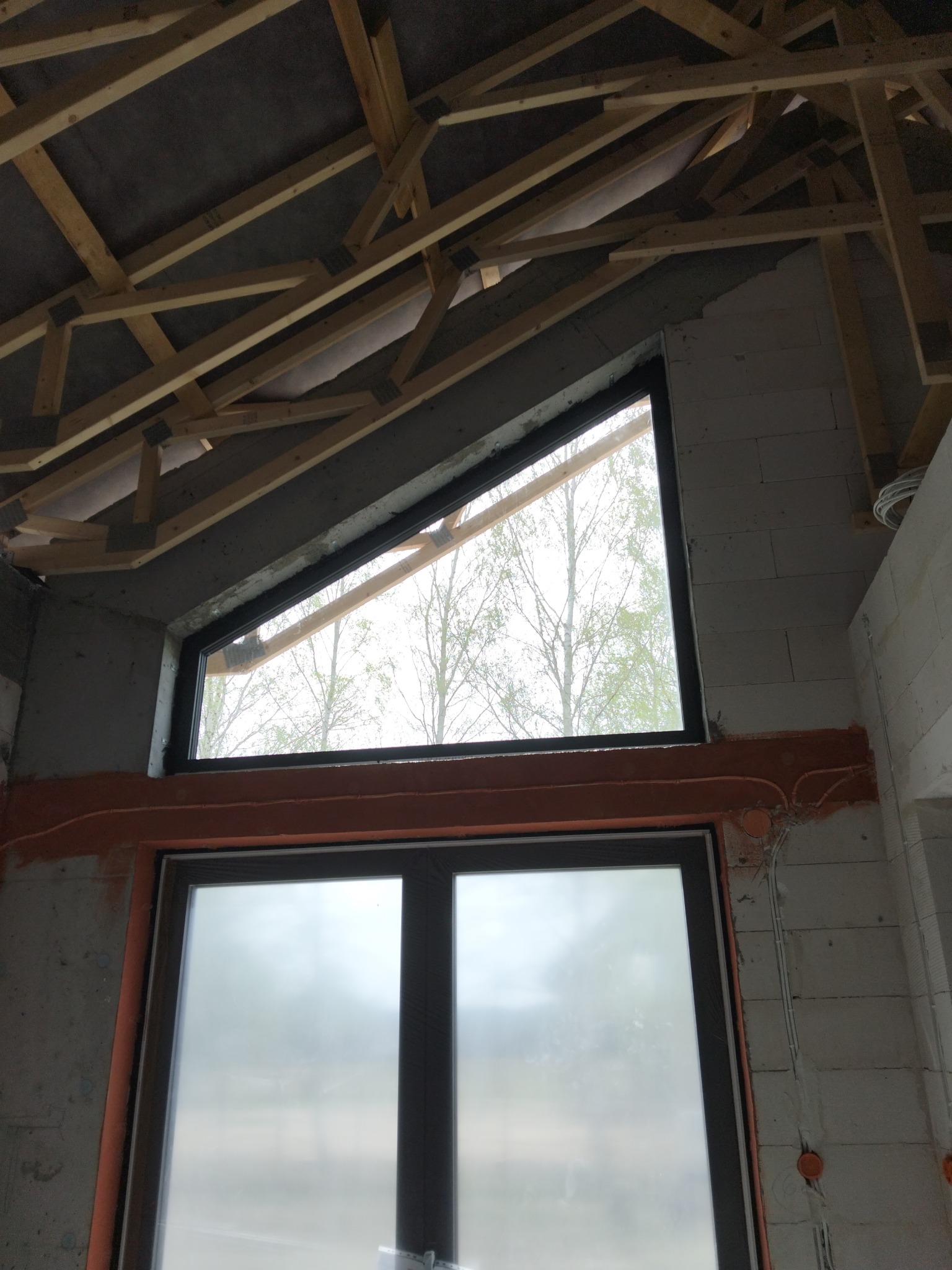 Wnętrze w stanie surowym z oknami: trapezowym u góry i prostokątnym na dole. Widoczne drewniane elementy konstrukcji dachu, instalacja elektryczna w ścianie z betonu komórkowego.