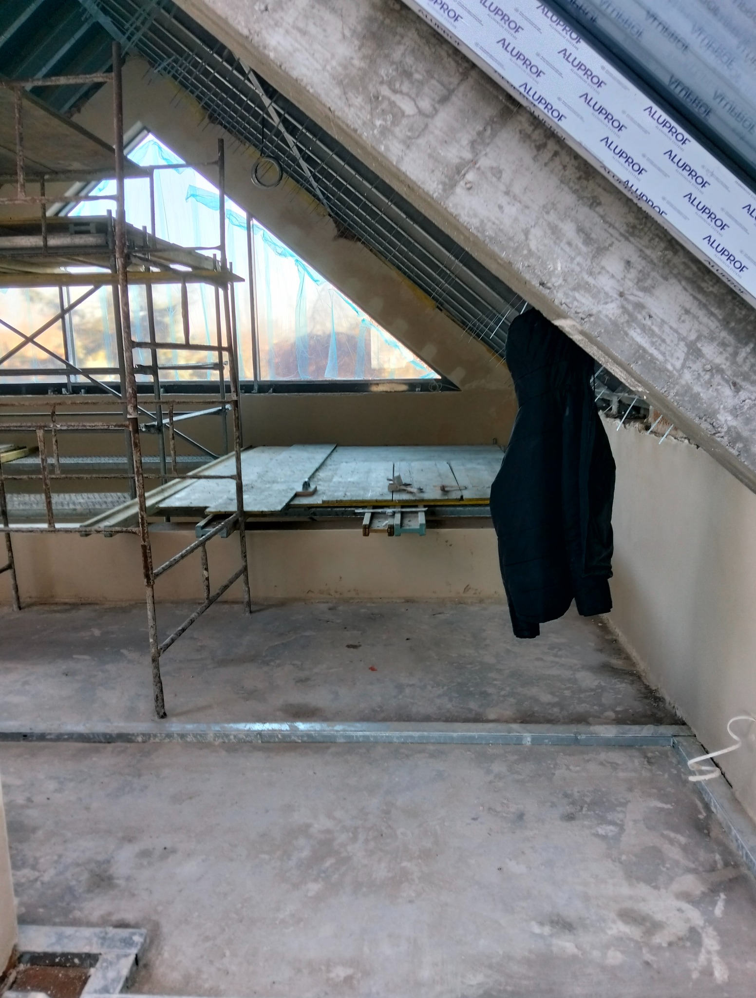 Skośne poddasze w trakcie prac wykończeniowych: rusztowanie, płyty na podłodze, wisząca kurtka. Widoczne profile sufitowe i okno dachowe.