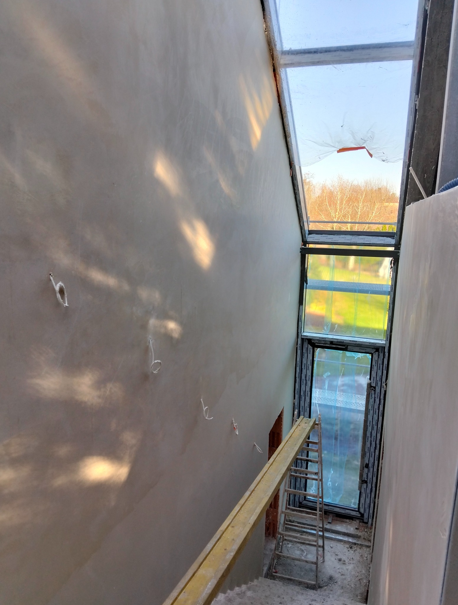 Ściana po szpachlowaniu w klatce schodowej z oknem dachowym, widok z góry. Widoczne ślady po przewodach elektrycznych i drabina. Wpadające światło słoneczne.