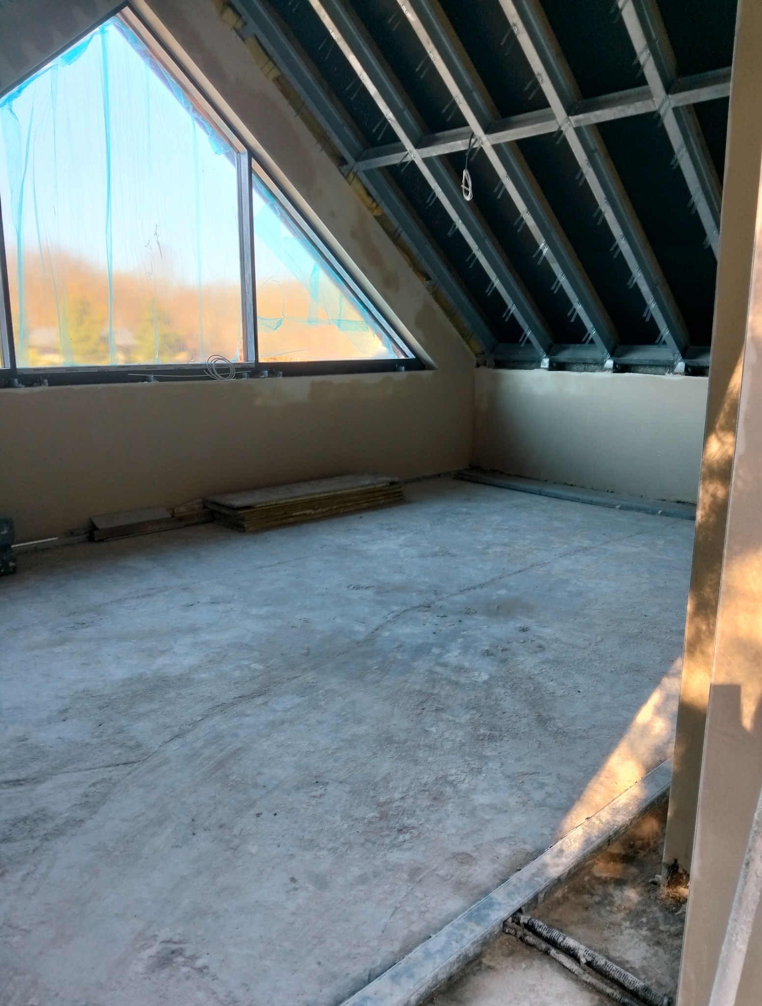 Poddasze w trakcie prac wykończeniowych: widoczne płyty karton-gips na ścianach, metalowa konstrukcja sufitu i okno dachowe zabezpieczone folią. Betonowa podłoga.