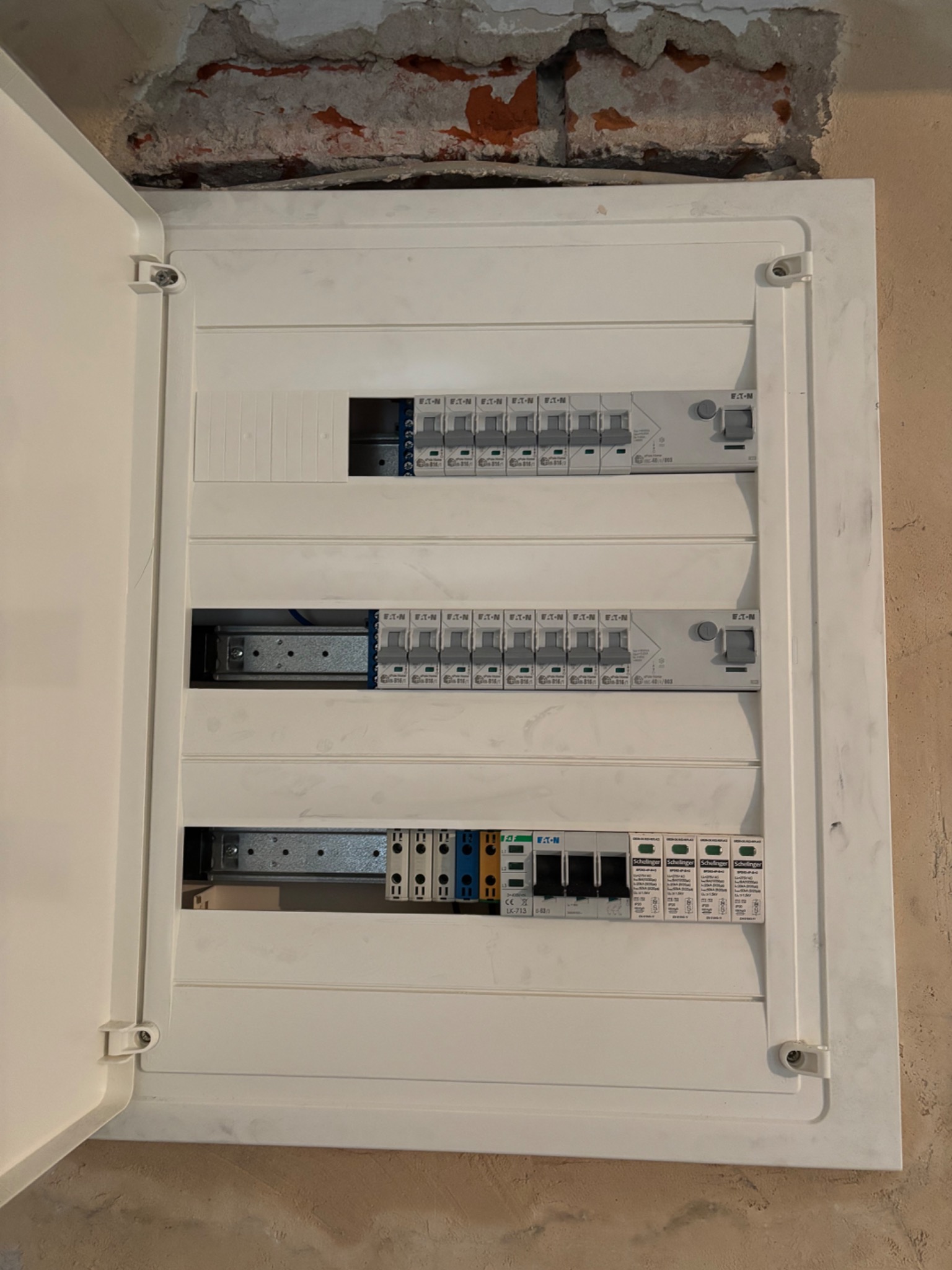 Otwarta, biała skrzynka rozdzielcza Eaton z zainstalowanymi bezpiecznikami i zabezpieczeniami, widoczna na tle surowej ściany podczas prac wykończeniowych.