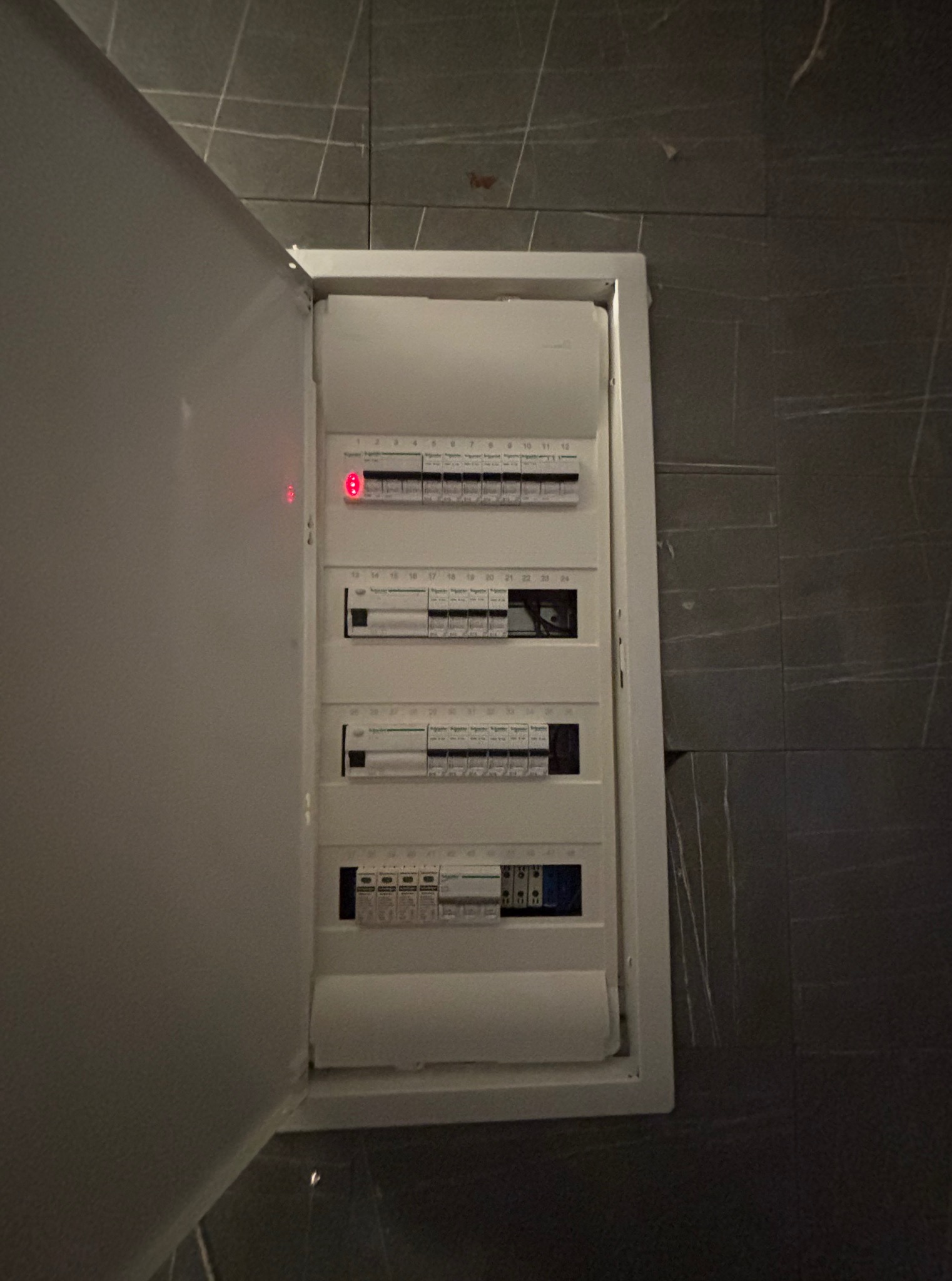 Otwarta skrzynka elektryczna z bezpiecznikami na szarej ścianie. Widoczne numery bezpieczników i oznaczenia producenta. Jeden z bezpieczników oznaczony czerwoną diodą.