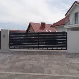 Nowoczesna, pozioma brama wjazdowa w kolorze antracytu, osadzona pomiędzy dwoma białymi słupkami. Widoczny podjazd z szarej kostki brukowej i dom z czerwonym dachem w tle.