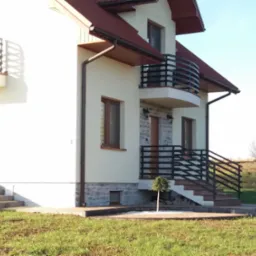 Nowoczesna balustrada balkonowa i schodowa z czarnych, metalowych profili przy domu jednorodzinnym w Ropczycach. Jasna elewacja, brązowe okna i dach.