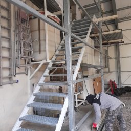 STEEL TEAM Strzałka Piotr - Metalowe schody przemysłowe z kratowymi stopniami, w trakcie montażu, z pracownikiem w odzieży roboczej. Widoczne elementy konstrukcyjne i drabiny w tle.
