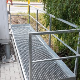 STEEL TEAM Strzałka Piotr - Metalowa kładka z kratą i barierką, łącząca budynek z terenem. Widoczne elementy konstrukcyjne, kostka brukowa i roślinność w tle. Szara kolorystyka.