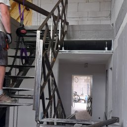 STEEL TEAM Strzałka Piotr - Montaż metalowej konstrukcji schodów w surowym wnętrzu. Widoczne elementy mocujące, pracownik w tle i dalszy plan z otwartym przejściem.