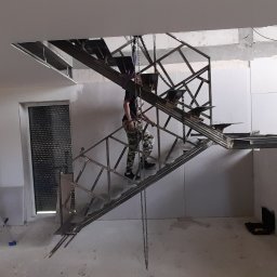 STEEL TEAM Strzałka Piotr - Montaż metalowych schodów w surowym wnętrzu. Konstrukcja podwieszona na łańcuchach, widoczny pracownik w trakcie instalacji. W tle okna z żaluzjami.