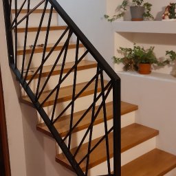 STEEL TEAM Strzałka Piotr - Nowoczesne schody z drewnianymi stopniami i czarną, geometryczną balustradą. W tle białe ściany z półkami i roślinami doniczkowymi. Ujęcie z dołu.