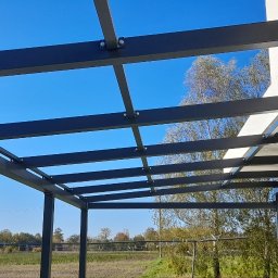 STEEL TEAM Strzałka Piotr - Nowoczesna, grafitowa pergola z metalową konstrukcją, widok z dołu na tle błękitnego nieba i zieleni. Konstrukcja przymocowana do budynku.