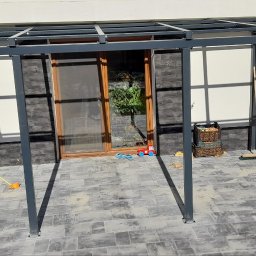 STEEL TEAM Strzałka Piotr - Metalowa konstrukcja zadaszenia tarasu w trakcie montażu, widoczne słupy i elementy mocujące, kostka brukowa na podłożu.